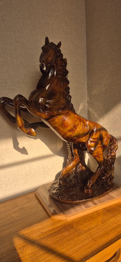 This media file features a detailed wooden sculpture of a horse in mid-gallop. The horse is depicted with its legs extended, mane flowing, and head turned as if in motion. The sculpture is carved from dark wood and has a polished, glossy finish, with some areas exhibiting a lighter, reddish-brown hue that suggests wood grain or a staining technique. It appears to be mounted on a textured base, possibly representing rocks or earth.
The sculpture is positioned against a textured, beige wall. Sunlight streams in from the right, casting distinct shadows of the horse and possibly a window frame onto the wall and the wooden surface to the right. The lighting suggests it is daytime, with the warm glow indicating either morning or afternoon sun. The wooden surface to the right, which appears to be a piece of furniture or paneling, has a visible wood grain pattern and a warm, reddish-brown color.
The scene is static, focusing on the artistry of the sculpture and the interplay of light and shadow. There is no indication of people or any specific event. The overall mood is one of quiet admiration for the craftsmanship. The location context of Gavar, Armenia, does not provide any specific visual cues within the image itself. No text is visible in the media file.