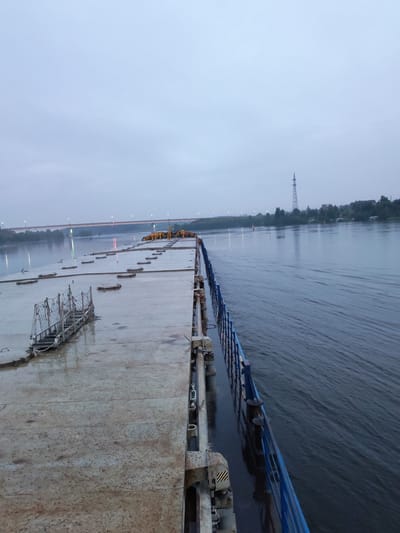 A wide, flat-topped barge or pontoon is shown on a calm river. The deck of the barge is made of concrete and appears wet, with some debris. A metal gangway with railings leads onto the deck from the left side. The barge has a blue railing along its right side, separating it from the river water, which is dark and shows ripples. In the distance, a long bridge with lights spans the river, and on the far bank, trees and a tall electrical pylon are visible. The sky is overcast and a pale blue-grey. The image seems to have been taken at dusk or on a very cloudy day. The location is identified as Ivanov Bor, Russia.