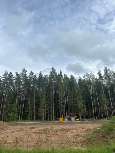 The image depicts a construction or landscaping project on the outskirts of a forested area. A red tractor with yellow attachments is positioned on a dirt road adjacent to a dense forest of tall pine and birch trees. The ground in the foreground is disturbed earth, with piles of soil and scattered stones. The sky is overcast with a mix of light and dark clouds. The scene suggests an ongoing outdoor work operation in a rural or semi-rural location within the Плешчаніцкі сельскі Савет, Belarus. There are no people visible in the image.