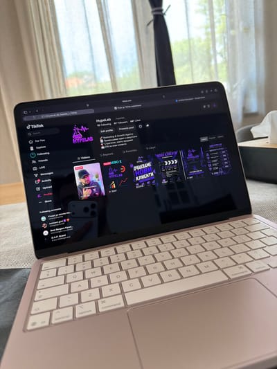 An overhead, slightly angled shot shows a pink MacBook Air laptop open on a gray couch with a textured fabric. The laptop screen displays the TikTok interface with a user profile for "HypeLab." The profile shows 10 followers and 47 following. The screen also displays several video thumbnails, some with Bulgarian text. The right side of the laptop screen shows a Google Docs document titled "Untitled document." A window with sheer white curtains is visible in the background, with hints of green foliage and a bright sky, suggesting an outdoor view. The scene does not provide any information about the city of Lozno, Bulgaria.