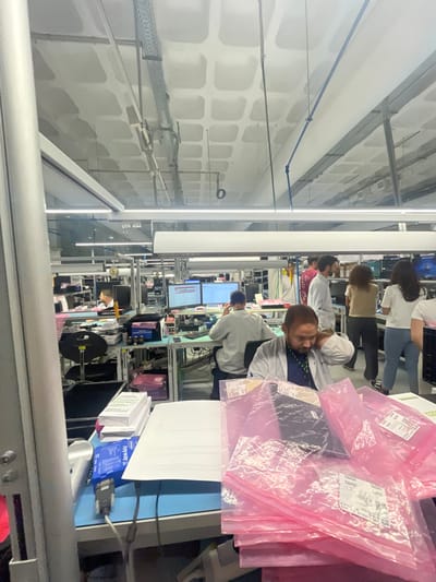 This image depicts an active open-plan workspace or laboratory in Amadora, Portugal. The environment features a concrete ceiling with a waffle-slab design, exposed metal cable trays, and long linear fluorescent light fixtures providing illumination.

In the immediate foreground, to the left, a metallic silver-gray vertical partition is visible. A light-blue tabletop extends across the lower portion of the frame, holding a stack of white papers and a blue "Nyval" electronic device with a DB9 serial port and attached cables. To the right, a substantial stack of translucent pink anti-static bags, each containing a dark, rectangular object and bearing white labels with text and barcodes, occupies the tabletop.

A male individual, with a dark beard and short hair, is seated behind this table, wearing a white lab coat over a dark blue lanyard. He is looking downwards and slightly to his left, with his right hand positioned near his ear or neck.

In the midground, multiple workstations are arranged in rows. One male, viewed from behind, is seated at a workstation, focused on two active computer monitors displaying data on dark backgrounds. He wears a light-colored long-sleeved garment. To his right, a group of four individuals stands, engaged in conversation. This group includes a bearded male in a light-colored, patterned shirt, another male in a light shirt, a female in a beige top and dark trousers, and another female in a white t-shirt and light blue jeans.

Further in the background, more workstations, computer equipment, and additional personnel are visible, indicating a busy operational setting. The overall scene suggests an environment focused on electronic assembly, testing, or similar technical work.