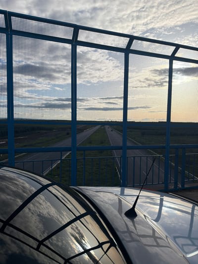 The image depicts an overhead view from a pedestrian bridge or overpass in Ait Boubidmane, Morocco, likely during sunrise or sunset. In the immediate foreground, the reflective silver roof of a vehicle, possibly a car, is visible, featuring a dark antenna and reflecting the cloudy sky and the structural elements above it. The midground is dominated by a blue metal grid fence, part of the bridge's safety barrier, which extends across the frame and partially obscures the view. Beyond the fence, a straight multi-lane asphalt road or highway stretches into the distance, flanked by green and brown fields under a wide, cloudy sky displaying hues of blue, grey, and orange from the sun's low angle. Two small, dark vehicles are faintly visible on the distant road. No people are present in the scene.