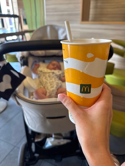 A hand is holding a McDonald's cup with a straw, angled downwards. The cup has a yellow and white striped pattern and condensation on the outside. Inside the cup, a baby is visible, lying in a stroller. The baby is wearing a patterned outfit and appears to be sleeping or resting. The background shows a blurry indoor environment with what appears to be green seating and wooden elements, suggesting a restaurant or cafe setting. There is also a glimpse of a stroller frame and a black object with a white shape on it in the foreground. The overall scene suggests a casual outing with a baby. The visible text on the cup is "0.5L" and the McDonald's logo. The time of day and weather are not discernible from the image.