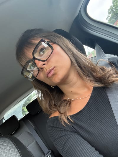 A woman with long brown hair and wearing black-rimmed glasses is in a car. She is wearing a black ribbed top and a necklace with small pearls. She has a seatbelt on. The car's interior is visible, with a gray seat and a window showing trees outside. The location is not discernible as being Saint-Laurent-du-Var, France based solely on this image.