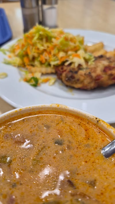 In Preiļi, Latvia, a close-up shot focuses on a bowl of creamy, orange-hued soup in the foreground, with visible specks of seasoning and subtle hints of ingredients. A silver spoon rests on the right edge of the bowl, partially submerged. In the background, a white plate holds a meal consisting of a piece of fried chicken or fish, a generous portion of coleslaw with shredded carrots and cabbage, and some french fries. The dining environment appears casual, with salt and pepper shakers visible on a table in the blurred background, suggesting a restaurant or eatery setting.
