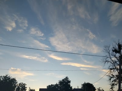 The image depicts a serene outdoor scene at dusk, likely in Cîrpești, Moldova. The sky dominates the frame, transitioning from a soft blue to warmer hues near the horizon, illuminated by the setting sun. Wispy, elongated clouds stretch across the sky, catching the fading light and adding texture to the celestial canvas. A thin black power line bisects the upper portion of the image, and another parallel line runs lower, subtly dividing the sky.

Silhouetted against the sky are the dark, indistinct shapes of trees and parts of buildings at the bottom of the frame. The trees are varied in density, with some appearing lush and full, while others are bare branches against the light, suggesting a mix of evergreen and deciduous types, or perhaps different stages of growth. A utility pole with attached wires is visible on the right side, further indicating a residential or semi-rural setting.

The overall mood is peaceful and tranquil, evoking the quiet transition from day to night. The lighting suggests it is either early morning or late evening, with the soft glow of the sun casting long shadows and bathing the clouds in a gentle light. There are no visible people or specific activities taking place; the focus is on the natural beauty of the sky and the subtle human presence suggested by the infrastructure. The scene is characterized by its simplicity and the quiet atmosphere of twilight.