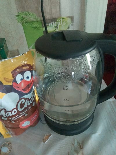 A close-up, slightly blurry, vertical shot shows a glass electric kettle filled with clear water, with condensation on the inside walls, placed on a textured surface. The kettle has a black lid and handle. To the left of the kettle is a yellow and brown packaged product with a cartoon penguin on it and text in Russian. In the background, a wall with a wood-grain pattern and a green rectangular object with the word "mojito" visible are present. The scene appears to be indoors. No individuals are visible in the image. The location is identified as Изыхские Копи, Russia.