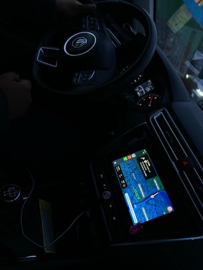 The image captures the interior of a vehicle from a high-angle perspective, facing forward towards the dashboard and windshield. The scene is dimly lit, suggesting dusk or night conditions.
Two hands belonging to a driver are visible: one is gripping the black multi-function steering wheel, which features a prominent chrome MG logo at its center and illuminated control buttons. The other hand rests near the manual gear shift lever, which displays a "1 2 3 4 5 R" pattern.
The dashboard's instrument cluster, partially obscured by the steering wheel, displays a digital reading of "136" (likely km/h) for speed, a fuel gauge, and the time "1:01".
A central infotainment touchscreen is active, displaying a navigation application, consistent with Apple Maps, featuring a blue route line. The map indicates the vehicle's current position on "Carretera Federal Mexico-Puebla." Visible points of interest and road names include "Calle Niños Heroes," "Calle San José," "Calle Leona Vicario," and a route segment leading to "Carrera al Aeropuerto Hnos Serdan" (Road to Hermanos Serdán Airport). The screen also shows the time "9:41" in its upper left corner. A small, pink peace-sign shaped charm is attached to the bottom edge of the screen.
To the right of the infotainment screen are vertical air vents, separated by a red hazard warning light button. A white charging cable is connected to a port below the screen, extending towards a compartment where a rectangular yellow-white card or paper is visible.
Through the windshield, blurred reflections of brightly colored objects and indistinct light sources are visible, suggesting an external urban or semi-urban setting.
Based on the navigation system's explicit display of "Carretera Federal Mexico-Puebla" and "Carrera al Aeropuerto Hnos Serdan," the vehicle is located on Federal Highway Mexico-Puebla, within the approximate vicinity of Santa María Zacatepec, Puebla, Mexico, and is actively navigating towards the Hermanos Serdán International Airport.