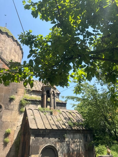 In Bjni, Armenia, a stone bell tower stands next to an ancient stone building with a domed roof. Green leaves from a nearby tree overhang the scene, casting shadows on the stone structures. The sky is clear and blue. The architecture suggests a historical or religious site, likely a church or monastery. A small bird is visible in the sky near a distant, smaller structure. A person is visible in the lower right corner of the image, sitting on a stone ledge.
