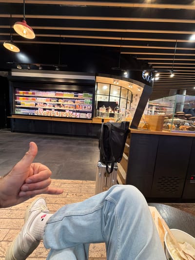 This image captures an indoor commercial food service establishment, likely within Lyon-Saint Exupéry Airport in Colombier-Saugnieu, France. The perspective is from a seated individual.
In the foreground, the lower body of one person is visible. This individual is wearing light blue denim jeans, white athletic shoes, and white ribbed socks with a small red design on the left sock. A left hand, positioned on the left side of the frame, performs a "shaka" or "hang loose" gesture. Immediately to the right of the person's legs, a silver, hard-shell suitcase is visible, topped with a black backpack. A partial view of a dark, round table in the bottom right reveals a white bowl containing food and wooden cutlery.
The background shows a contemporary cafeteria or market-style setup. On the left, a large, refrigerated display unit against a black wall is fully stocked with various pre-packaged beverages and food items arranged on multiple shelves. To its right, a curved counter with a dark base and a lighter wooden top extends across the midground. Partially visible signage above this counter reads "EAKI". Behind this counter, a glass display case presents baked goods, and other food service elements, including silver-domed food warmers, are present. The wall behind the counter features a grid-patterned design.
The ceiling is dark, intersected by numerous horizontal light-colored wooden slats. Several light fixtures are visible, including two red, bell-shaped pendant lights on the upper left and track lighting. The floor transitions from a light brown, possibly wood-patterned, surface in the foreground to a darker grey floor further into the establishment. The scene is brightly illuminated by artificial lighting.