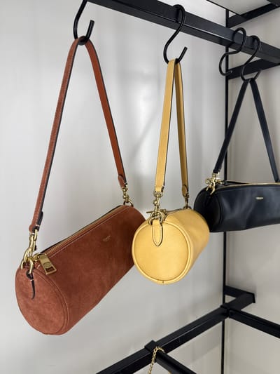 The image shows three cylindrical Coach handbags hanging from a black metal rack. The bags are arranged vertically, one above the other. The top bag is black leather, the middle bag is yellow, and the bottom bag is brown suede. The rack is located in Sunrise, United States of America.