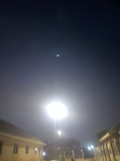 The image, taken at night in Ebo, Nigeria, shows a street lit by a bright street lamp. A building with multiple windows is visible on the right side of the frame. The sky is dark, with some ambient light visible.
