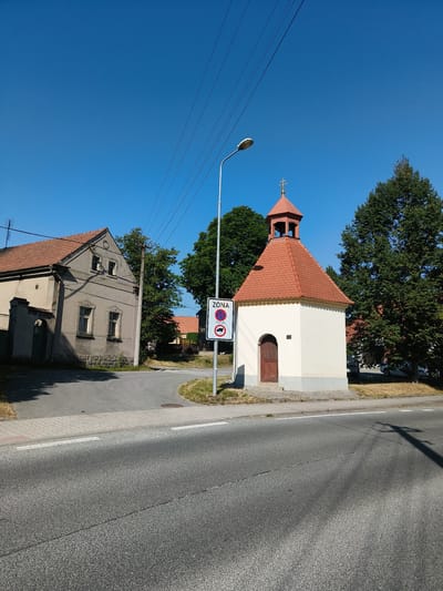 The image depicts a rural scene in Útušice, Czechia. In the foreground, a paved road curves to the right. Adjacent to the road, a sidewalk made of interlocking concrete pavers is visible. A small, cream-colored chapel with a red tiled roof stands to the right of the road. The chapel has a single wooden door and a small bell tower topped with a cross. To the left of the chapel, a street sign with a red circle and a diagonal bar, indicating a prohibition, is mounted on a pole. Below this symbol is another symbol of a truck, also within a red prohibition circle. A lamppost stands behind the sign.
Behind the chapel, a large green tree is present. Further back and to the left of the chapel, a gray building with a red tiled roof is visible. This building has multiple windows and a sloped roof. A dark green fence and gate are positioned in front of this building. Power lines stretch across the blue sky above the scene. The environment appears to be dry, with patches of dry grass and dirt along the side of the road and around the buildings. The sky is clear and bright blue, indicating a sunny day.