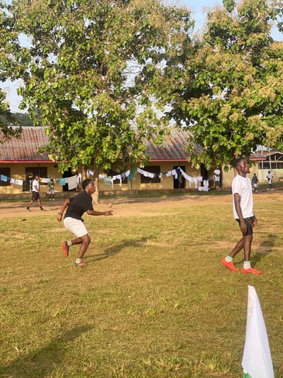 In Ise- Ekiti, Nigeria, two young men are engaged in a friendly football match on a grassy field. One man, wearing a white t-shirt and black shorts with red soccer cleats, is captured mid-air, seemingly kicking a ball. His body is contorted in an athletic pose, suggesting a dynamic moment in the game. Beside him, another man in a black t-shirt and white shorts, also wearing orange soccer cleats, is also in motion, with one arm raised and the other extended towards the ground. His posture implies he might be dribbling or defending.
In the background, a building with a corrugated iron roof stands, with laundry hanging on a line in front of it, suggesting a residential or communal area. A large tree with green foliage and small blossoms is prominently featured on the left side of the frame, providing shade and framing the scene. The overall setting appears to be a sunny day, with the warm light casting shadows on the grass, indicating it is likely late afternoon. The atmosphere seems casual and energetic, with the focus on the sporting activity and the camaraderie between the participants. No visible text is present in the image.