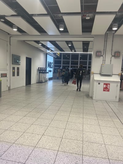 This media file captures an indoor scene, likely within an airport terminal or a large transit hub in Horley, United Kingdom. The perspective is from slightly behind a group of people, looking forward into a well-lit corridor or concourse.
The floor is covered with light-colored, speckled square tiles. On the right side of the image, a white "FIRE HOSE REEL" cabinet is prominently visible, with various electrical conduits and grey boxes mounted on the wall above it. The ceiling, running along the left side of the frame, features a grid of white rectangular panels interspersed with recessed circular lights, casting a bright illumination over the area.
In the mid-ground, a small group of people are walking or waiting. Most are seen from behind or the side. The most distinct individual, positioned slightly ahead of the others, wears a dark, form-fitting athletic top with "IRON MAN" clearly visible on the back, and carries a red bag in their left hand. Other individuals are dressed casually and appear to have bags or luggage, consistent with travel.
Further into the background, a large, illuminated sign displays "M&S Food," indicating a Marks & Spencer food outlet. Beyond the people and the sign, large windows suggest an exterior area, and the darkness visible through them, combined with the reliance on artificial lighting indoors, points to the evening or nighttime. The overall atmosphere appears neutral and functional, typical of a busy public transportation area.