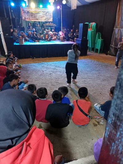 This nighttime image captures a vibrant cultural performance taking place in Kecamatan Cimaung, Indonesia, likely under a large tent or in an outdoor venue. An elevated stage, illuminated with bright blue and purple lights, features a group of approximately 10-15 male musicians. They are seated or standing, playing various traditional Indonesian instruments, including drums (kendang) and gongs. A banner hangs at the back of the stage, displaying partially visible text that reads "ANGKLUNG SAHA SAUNGA REAK CI... MANGSA," indicating the type of traditional music or performance.

In front of the stage, on a dirt ground, a woman seen from the back is performing or dancing barefoot, wearing a grey long-sleeved top and dark pants. To her right, another individual is also visible, barefoot, seemingly engaging in the performance. A large audience, primarily consisting of children and young boys, sits on the ground in front of the stage, attentively watching the event. Their faces, when visible, show engagement, while others are seen from behind, their focus directed towards the stage. In the immediate foreground, a person in a red hoodie with a black head covering is also part of the audience. To the right of the stage, several green plastic chairs are stacked, and large speakers stand on either side of the stage. The overall scene depicts a lively community gathering centered around traditional music and dance.