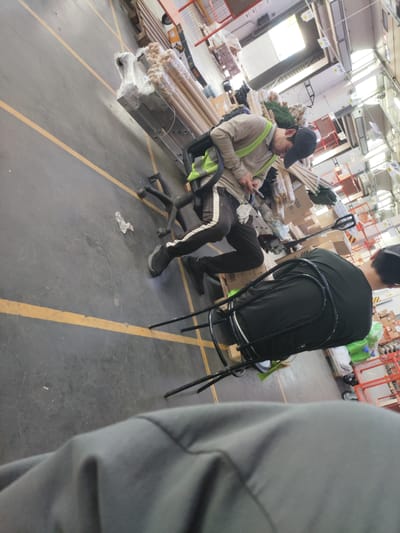 The image depicts an interior scene of a warehouse or retail store, possibly a hardware or home improvement store, with a concrete floor marked with yellow lines. The perspective is from a low angle, looking upwards and to the side. In the mid-ground, a person wearing a light brown long-sleeved shirt, a green reflective vest, dark pants with white stripes, and a black baseball cap is partially visible. This person appears to be operating a wheeled mechanism, possibly a cart or a lift, and is holding a tool or a small object in their hands. To the right and further back, another person, wearing a dark green t-shirt and dark pants, is seated or leaning on a black wheeled chair. This individual is wearing a black baseball cap and is facing away from the camera. The environment is filled with various objects, including stacks of merchandise, pallets, wooden poles wrapped in plastic, and shelving units in the background. The lighting suggests an indoor space with overhead illumination, possibly from skylights. The overall impression is a working or storage area. The location is identified as Potapovo, Russia.