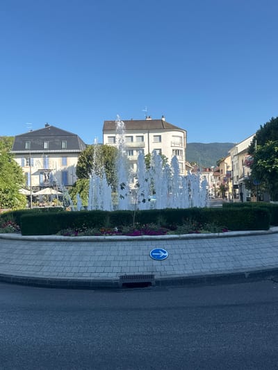 This image shows a public fountain in a roundabout in Divonne-les-Bains, France. The fountain is centrally located, with water jets spraying upwards. Around the fountain is a circular garden bed filled with colorful flowers and green bushes. The roundabout is paved with light-colored interlocking stones, and a blue traffic sign with a white arrow indicating a left turn is visible on the pavement. Beyond the roundabout, the scene depicts a street lined with multi-story buildings, some with balconies. Trees and hills are visible in the background under a clear blue sky. The overall impression is of a pleasant, well-maintained urban square.