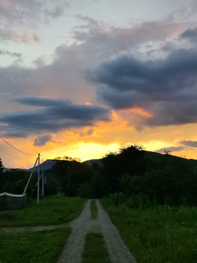 The image depicts a rural landscape at dusk, likely in Bagovskaya, Russia. A dirt road stretches into the foreground, leading towards a line of dark trees and hills in the distance. The sky is filled with dramatic clouds, illuminated by the warm, orange and yellow hues of the setting sun, contrasting with the darker, grey clouds above. Power lines and poles are visible on the left side of the road, suggesting the presence of rural infrastructure. A small, dark structure, possibly a shed or outbuilding, is situated near the power lines. The ground on either side of the road is covered in green grass, with some wildflowers visible in the denser patches. No people or animals are present in the scene.
