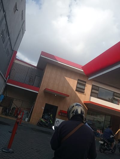 This media file shows an outdoor scene at what appears to be a gas station or convenience store in Surabaya, East Java. The perspective is from the back of a person wearing a helmet, looking towards a building with a modern design featuring red and tan elements. The sky is partly cloudy, suggesting it might be late afternoon or early morning.

The building has large windows, some of which are dark and reflective, showing a hint of the interior. There are also some smaller, louvered windows. Red accents are prominent on the roofline and some of the building's trim. In the foreground, red bollards are placed on a paved surface made of interlocking bricks.

Several motorcycles are visible, indicating a common mode of transport in the area. One green motorcycle is parked near the entrance of the building, and another is being ridden by a person in the mid-ground. Further in the background, another person is on a motorcycle.

Visible text in the image includes "Mas" on the helmet of the person in the foreground, and signage on the building that reads "AIR RADIATOR ISI ANGIN BAN" (Air Radiator, Tire Inflation) and "JUAL AQUA SGALIVON" (Selling Aqua Gallons). License plates are also partially visible on the motorcycles. The overall impression is a busy, everyday scene at a commercial establishment.