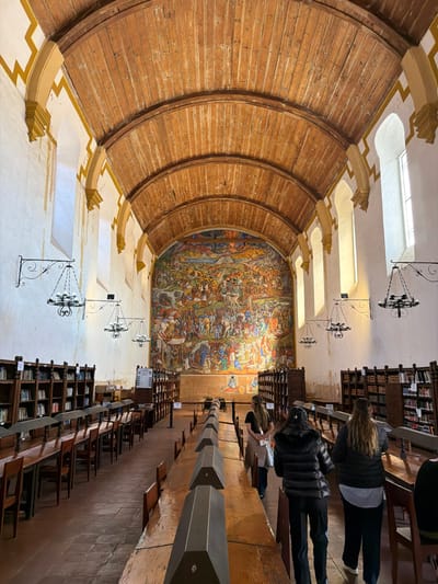The image displays a grand, historic interior space, likely a library, archive, or a significant hall within a public building, illuminated by natural light during the daytime. The perspective is tilted, but describing it as if oriented correctly:
To the left, a large, impressive curved wall or ceiling section dominates the space, constructed from numerous horizontal wooden planks that create a deep, ribbed, and visually compelling arc. Beneath this wooden structure, a light-colored wall is decorated with ornate, possibly gilded or yellow-painted, geometric patterns, featuring prominent corbels that jut out, providing structural or decorative support to the wooden curve above. High on this wall, several rectangular windows or skylights allow ambient light to flood the room.
Across the room, on what appears to be the far wall, a vast and vibrant mural is the focal point. This colorful artwork depicts a complex, densely populated scene, possibly historical or allegorical, featuring numerous figures engaged in battles, processions, or other significant events. The mural's palette includes rich earth tones, blues, and reds, contributing to its dramatic impact.
From the ceiling, several ornate black wrought-iron chandeliers hang, adding a touch of antique elegance to the setting.
On the right side of the image, multiple rows of tall, dark wooden bookshelves are visible, neatly filled with numerous books, indicating an extensive collection. In front of these bookshelves, long, sturdy wooden tables or benches extend into the distance. Several individuals, mostly seen from behind or in partial profile, are present among these tables and bookshelves. They appear to be adults, dressed in casual attire, including jackets and pants, suggesting they are either browsing the collection, studying, or observing the magnificent mural and the architecture of the hall. Their postures suggest calm engagement with their surroundings.
The overall atmosphere conveys a sense of historical significance, scholarly pursuit, and artistic beauty. No legible text is visible within the image.