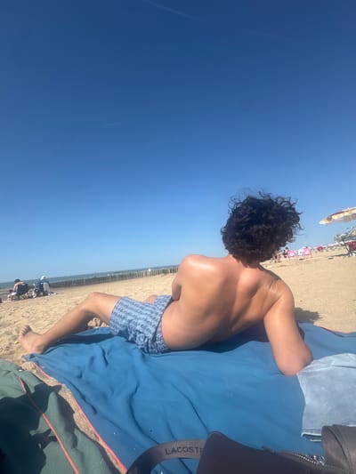 A man is lying on a blue towel on a beach. He is wearing blue patterned swim trunks. The towel is partially on top of a dark green blanket. To the right is a black bag with "LACOSTE" visible on the strap. People are seated on the sand in the distance with beach umbrellas. The sky is clear and blue. The location is a beach in Knokke-Heist, Belgium, evidenced by the line of wooden breakwaters.