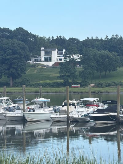 The image shows a body of water with several boats docked. In the background, there is a large modern house on a hill surrounded by trees. The sky is clear and light blue. The boats are of various sizes and types, with some covered and others open. The water is calm, with reflections of the boats and the sky visible. The environment appears to be a marina or a waterfront residential area. The location is identified as the Village of Bayville, United States of America.