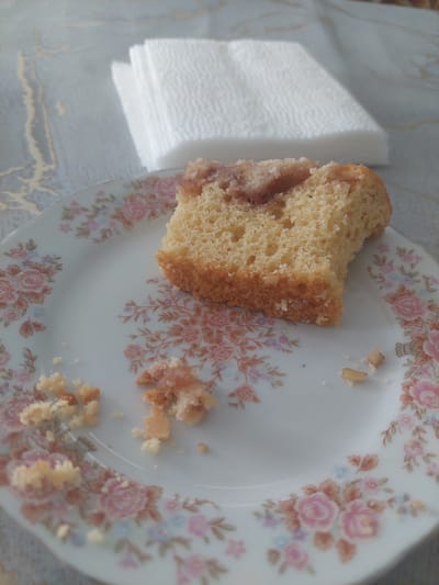 A slice of cake with a reddish swirl pattern is on a floral patterned plate. Crumbs are scattered around the plate. In the background, a stack of white paper napkins sits on a grey and white marbled surface. The scene is likely indoors.