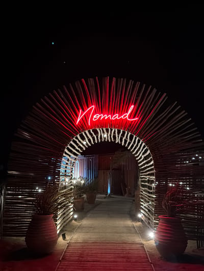 The image depicts an archway constructed from natural wood sticks, illuminated by red neon signage spelling out "Nomad." The archway is set against a dark night sky. A wooden walkway leads through the archway, flanked by large, textured terracotta pots containing sparse vegetation. Small, upward-facing spotlights are embedded along the walkway and the base of the archway, casting a warm glow. The environment appears to be a desert or sandy area, with the ground around the walkway being sandy. In the sky, two small, bright celestial bodies are visible. The overall scene suggests an entrance to an establishment or event, possibly a restaurant or bar, in a bohemian or rustic setting. The location is identified as the Nomad beach bar in Torreilles, France.