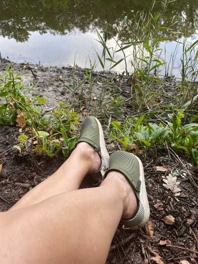 A first-person perspective image shows a person's legs and feet, wearing olive green Crocs, resting on the muddy bank of a body of water. The water reflects the sky and trees. Green reeds and dry leaves are scattered around the feet. The overall scene suggests a natural outdoor setting, possibly a park or waterfront area.
