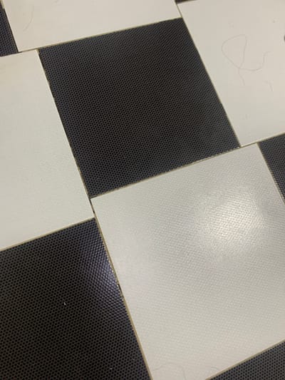 The image shows a close-up view of a black and white tiled floor, possibly in a home or public building. The tiles are arranged in a checkerboard pattern. Some of the white tiles have faint lines or stray hairs on their surface. The black tiles have a textured, dotted pattern. The lighting suggests an indoor environment, though no specific details of the city of Glodeni, Moldova are visible.