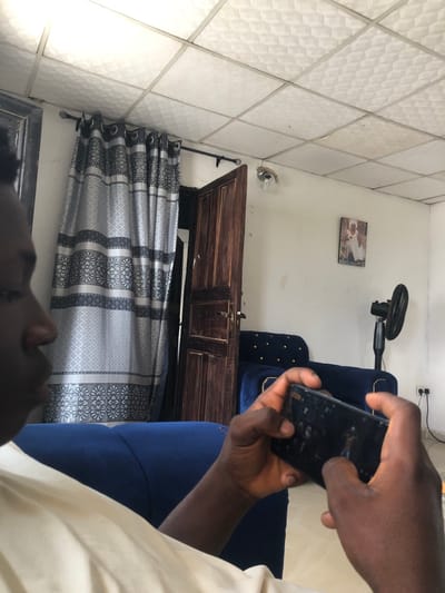 The image depicts an indoor scene within a residence in Abatete, Nigeria. A person, identified by dark skin, is seated on a dark blue, possibly velvet, sofa. The individual's left facial profile (nose, cheek, and chin) and upper torso, clad in a light-colored garment, are visible on the left side of the frame. Both hands grasp a black smartphone horizontally, with thumbs positioned on the screen, indicating active engagement with the device, which displays illuminated digital content.
The background features a window on the left covered by a grey and white patterned curtain, suspended from a black rod with decorative finials. Centrally, an open, dark brown paneled wooden door provides access to an adjacent area. The ceiling consists of white, square, textured suspended tiles. A simple light fixture with a bare bulb hangs from the ceiling near the doorframe. The white wall on the right displays a framed picture of an individual in white robes. A black standing electric fan is positioned on the floor against this wall, next to two white electrical outlets, one of which has a black power cord plugged in. A portion of the dark blue sofa, featuring visible stud detailing, is also present on the mid-right side of the image.