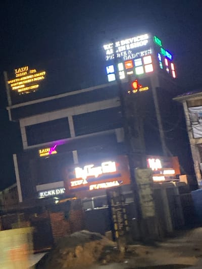 Nighttime view of a tall building adorned with illuminated signs. “KC E Devices Al In 1 Shop Ye Me & Gadgets” is stacked vertically. Below, signs for “Ladu Salon Spa” and “KCKE DE” can be discerned. An adjacent sign features a stylized text “HEDA” and what appears to be "SWAXHA." The lighting casts a vibrant glow, adding to the dynamic energy of the cityscape.