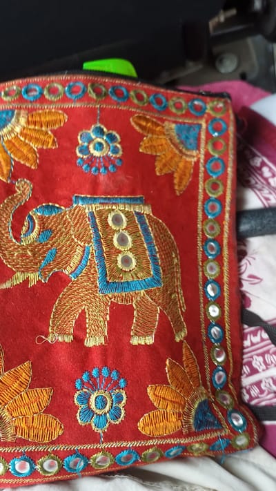 The image shows a close-up of a red embroidered fabric, likely a pouch or a decorative panel. The embroidery features an elephant in gold and blue thread, adorned with small, reflective circular embellishments on its howdah. The borders of the fabric are also decorated with intricate gold and blue stitching, interspersed with small, round, mirrored pieces. There are also embroidered floral motifs in blue and orange. The background is blurred and suggests an indoor setting, possibly a flat surface covered with patterned fabric. There is no indication of the city of Киясово, Russia, in the image or its details.