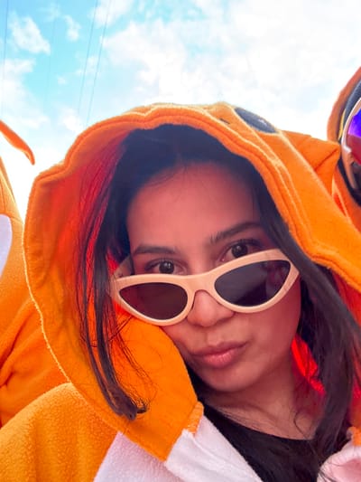 A person in an orange and white hooded animal onesie and white-framed sunglasses tilts their head towards the camera. The hoodie's design includes round black spots resembling eyes. Dark hair frames the face. The sky with wisps of clouds is visible in the background. A blurry object is seen above the head.