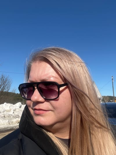 The image displays an outdoor scene in the city of Сундур, Russia, under a clear blue sky. An adult female is featured prominently in the foreground, positioned slightly off-center and looking towards the viewer's right with a subtle smile. She has long, straight, light-blonde hair and wears dark-framed sunglasses with dark, possibly reddish-purple, tinted lenses. Her attire includes a black hooded jacket over a dark undershirt. In the background to the left, a large, partially melted snowdrift is visible on the ground, accompanied by bare tree branches and a distant treeline on a hill. To the right, behind the woman, a dark-colored vehicle, a light-colored building, and a wooden utility pole with power lines are discernible. The scene is brightly illuminated by direct sunlight, and the presence of snow indicates a cold season, likely late winter or early spring.