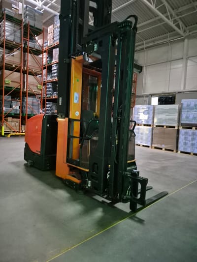 A forklift is parked in a warehouse with shelves stacked high with boxes. The forklift is orange and black. The warehouse has a concrete floor and a high ceiling with exposed metal beams. The shelves are metal and appear to be orange. There are several stacks of boxes on the shelves. The boxes are of various sizes and are wrapped in plastic. The scene is lit by overhead lights. The location is identified as Pabradė, Lithuania. There are no people visible in the image.