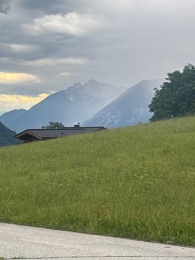 A wooden cabin with a dark, shingled roof and a chimney is situated on a grassy hillside. The cabin is partially obscured by the foreground, which is a vibrant green meadow dotted with small white flowers. In the background, majestic mountains rise, their peaks shrouded in mist and clouds. The sky is overcast, with patches of sunlight breaking through the clouds on the left side of the image, casting a golden glow. A paved path or road runs along the bottom edge of the frame. The scene evokes a sense of natural beauty and tranquility in a mountainous region. The location is Hyna, Austria.