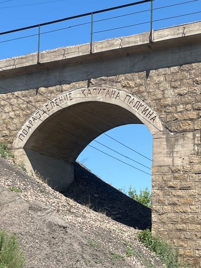 The image shows an arched underpass, likely part of a railway bridge, constructed with rough-hewn stone blocks on the exterior and a smooth concrete interior. Carved into the concrete arch is text in Cyrillic script, reading "ПОДРАЗДЕЛЕНИЕ КАПИТАНА ПАЛКИНА". This translates to "Captain Palkin's Unit". The underpass is situated on a slope covered with loose gravel and vegetation, with power lines visible above the arch and against a clear blue sky. The overall scene suggests an infrastructure element in a somewhat rural or industrial setting. The provided information about the city of Aqtau, Kazakhstan, is not directly verifiable from the visual elements alone.