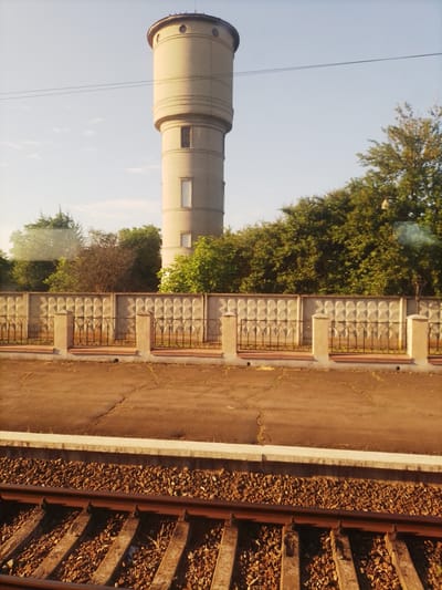 An outdoor, slightly hazy, horizontal medium shot captures a water tower and a train track in Pokrovske, Ukraine. The water tower, made of light-colored bricks, has a cylindrical shape with several distinct bands and two small rectangular windows. It is positioned on the left side of the image and rises above a dense line of green trees. In front of the trees, a concrete fence with a repeating diamond pattern is visible, supported by concrete pillars and a black metal railing. A cracked asphalt surface is in front of the fence. The foreground is dominated by a section of train tracks, with rusty metal rails resting on wooden sleepers embedded in gravel. The sky is a pale blue with a few scattered white clouds.