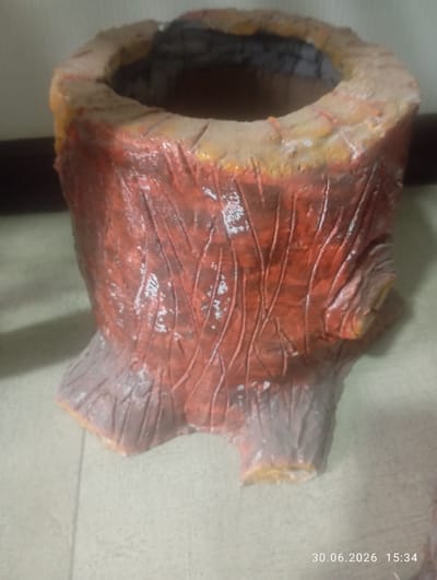 A close-up, medium shot captures a decorative pot shaped like a tree stump. The pot is made of a rough, textured material, painted in shades of reddish-brown and light grey to mimic the bark and base of a tree. Vertical, carved lines suggest the natural grain of wood. The top opening of the pot is visible, revealing a dark, hollow interior. The pot is positioned on a light-colored, tiled surface. The timestamp "30.06.2026 15:34" is overlaid in the bottom right corner of the image. There are no people or animals present in the scene. The background is a plain, light-colored wall and a dark, horizontal surface, suggesting an indoor setting. The lighting is soft and diffused. No specific landmarks or elements of Ohangaron, Uzbekistan, are discernible within the frame of the image.