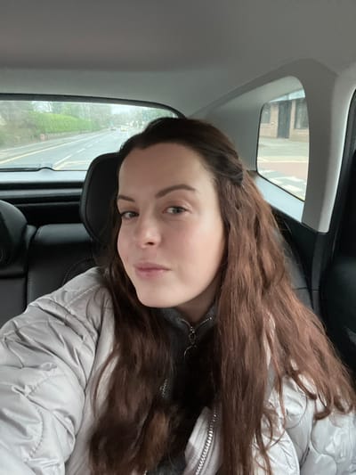 A female subject, appearing to be in her 20s or 30s, with long, wavy, dark brown hair and light skin, is depicted from the chest up, seated in the rear passenger compartment of a vehicle. She wears a light grey quilted puffer jacket over a dark grey zipped top with a ring-pull zipper and is looking directly towards the camera with a neutral expression, a slight upturn visible at the corners of her mouth.
The vehicle's interior shows dark-colored seating and a light-colored ceiling. Through the rear window, a two-lane road is visible, marked with white lines, including a solid line on the left edge and a dashed central lane divider. A long, dense green hedge and trees line the left side of the road, which shows distant vehicles. Through the right rear passenger window, a two-story, dark brick building with dark rectangular openings (likely windows or doors) is observed. A paved area with white horizontal markings, consistent with a pedestrian crossing, and a yellow curb line are visible between the building and the road. The scene is illuminated by ambient, overcast daylight. The specific street or town within the district of South Oxfordshire, United Kingdom, cannot be definitively identified from the available visual information.