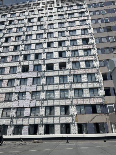 The image depicts a multi-story building under renovation. The facade is partially covered with white insulation material, some of which has a red star pattern. Metal framing is visible, suggesting an ongoing construction or cladding process. Numerous windows are present, some covered with plastic sheeting, while others appear open or incomplete. The building is adjacent to an older section of the structure, which shows signs of wear and tear, with exposed materials and discolored surfaces. The scene is viewed from a rooftop or elevated position, with a dark roof surface in the foreground featuring some coiled rope and electrical cables. The sky above is overcast and gray. The location is identified as Аснежыцкі сельскі Савет, Belarus.