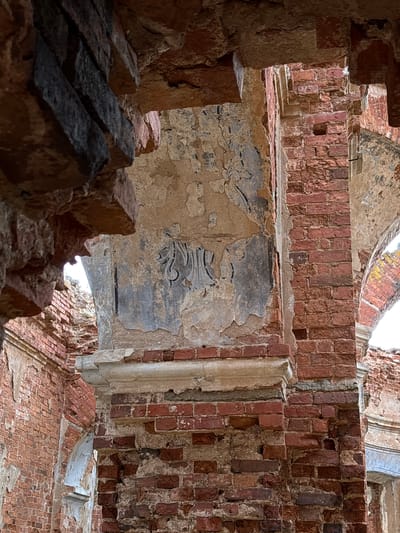 The image depicts the interior ruins of a building in Staroe Rakomo, Russia. The structure is primarily constructed of red brick, with significant sections decayed and crumbling. Plaster is visible on some wall surfaces, displaying remnants of decorative patterns in dark gray or black. The view is framed by sections of broken brick in the foreground, drawing the eye to the open spaces of the ruined structure. Light filters through gaps in the walls and ceiling.