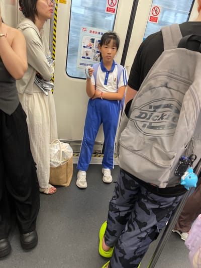 This is a photograph taken inside a subway car, likely in Longhua District, China. The image is oriented vertically. In the foreground, a young girl wearing a blue and white uniform is seated, holding what appears to be a small item in her hand. She has dark hair and is looking towards the camera with a neutral expression. Behind her, a person's back is visible, wearing a grey backpack with the "Dickie" logo prominently displayed. A small blue shark toy and a toy car are attached to the backpack.
Further down the subway car, two other people are seated. One individual wears glasses and a light-colored top, with their arms crossed. Another person is wearing a long, cream-colored skirt. Black pants and dark shoes are visible at the bottom of the frame, belonging to another passenger.
The setting is clearly a public transportation vehicle, indicated by the grab bars, the subway car window showing a blurred exterior, and the safety signs. One sign, written in Chinese, contains text like "禁止倚靠" (No leaning), "车门正在关上" (Doors are closing), and "请勿上/下车" (Do not get on/off). Another sign features a red circle with a diagonal line through it, signifying prohibition, along with text related to mosquito-borne diseases like dengue fever, mentioning symptoms like muscle aches and fever, and urging prevention through mosquito bites. The general atmosphere suggests a typical commute on a subway. The lighting appears to be artificial, consistent with an indoor environment. No specific time of day or weather conditions are discernible.