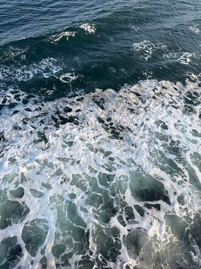 The image presents an overhead view of the sea surface in the city of Kato Karioutes, Greece. The scene is dominated by dark teal-blue water exhibiting varying degrees of agitation. In the foreground and midground, a substantial expanse of white, irregular foam covers the water, forming intricate, interconnected patterns and obscuring the underlying water in many areas. The foam gradually diminishes towards the upper portion of the frame, transitioning to deeper, less foamy water with visible wave crests and troughs. No individuals, vessels, or man-made objects are present. The lighting is consistent with daylight conditions, casting subtle illumination on the foam's texture. The water is in a state of dynamic movement, indicated by the wave formations and the widespread presence of aerated foam.