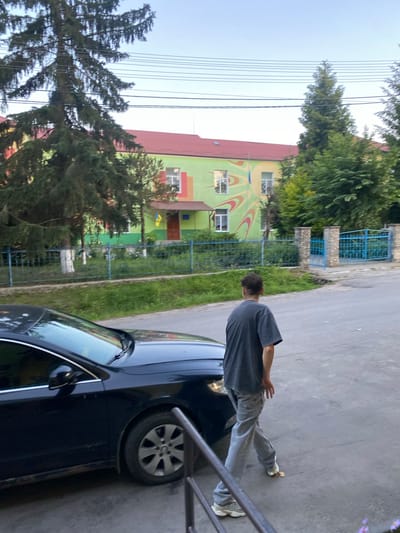 The image captures a street scene with a dark-colored sedan parked on the left side. A person, viewed from behind, is walking away from the camera towards the right, wearing a dark gray t-shirt and light-colored pants. Behind the car and the walking person, a building with a green facade and a red roof is visible. The building features windows and a mural with abstract yellow and orange designs. A blue metal fence runs along the front of the building, with a gate visible to the right. Trees and green grass are present in the area between the fence and the building. The sky above is clear. The scene appears to be in the city of Tovstoluh, Ukraine, as indicated by the presence of a Ukrainian flag flying near the entrance of the building. The overall atmosphere is that of an outdoor, possibly residential or institutional, setting.
