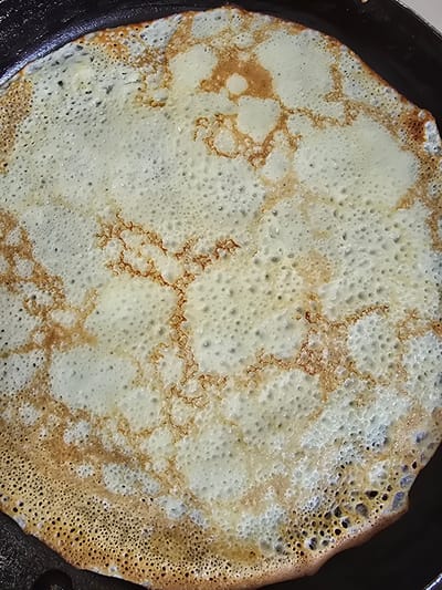 The image shows a close-up, overhead view of a single, thin crepe or pancake cooking in a black frying pan. The crepe is circular and fills most of the frame, with the dark rim of the pan visible around the edges. The crepe itself has a slightly uneven texture with many small bubbles and some areas of browning, indicating it is actively cooking. The browning is not uniform, with some parts appearing golden brown and lacy, while others are pale yellow. The crepe is cooked on one side, with the texture of the cooked batter clearly visible. There are no people or other objects visible in the image. The scene is purely focused on the cooking process of the crepe. There is no information or context provided in the image that would allow for its location in the city of Satbayev, Kazakhstan.