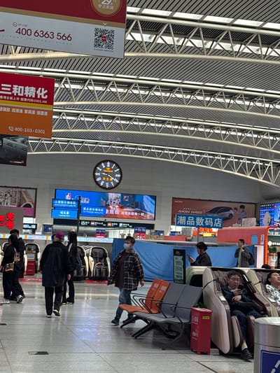 The photograph captures a busy interior scene, likely a train station or airport terminal in Huanggu District, China, judging by the visible signs and posters written in Chinese. The dominant features are the architectural elements: a high ceiling supported by a complex structure of steel beams and diagonal supports, creating a sense of spaciousness.

The scene is filled with people in varying activities. Some individuals are seated in massage chairs, while others are walking or standing around. A man in a plaid shirt and mask stands near the seating area. The crowd of people are dressed in dark clothing, suggesting a cool or transitional season.

Numerous signs and advertisements adorn the walls, featuring Chinese characters and recognizable brand names. A large clock is mounted on the wall with “TIANWANG” written on it. Other signs include travel directions.

The scene also includes advertisements for products and services, a modern SUV, and promotional materials for businesses.