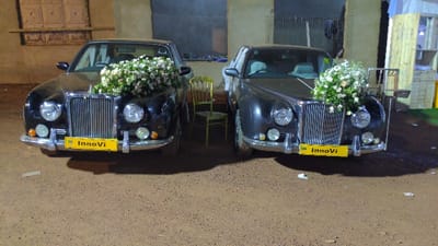 Two vintage black cars sit side-by-side, adorned with bouquets of white flowers. Each has a yellow license plate with "InnoVi" on it. A gold chair is placed between the cars. The ground appears to be a dirt lot. There are buildings in the background.